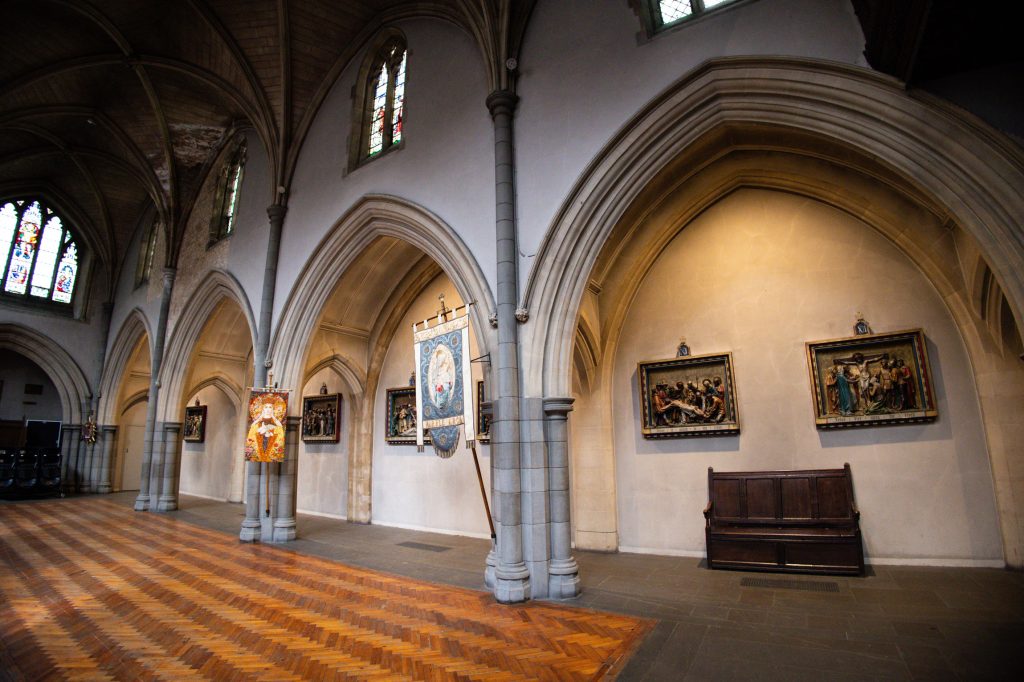 South aisle of the Church of the Annunciation with Gothic arches, devotional artwork and patterned wooden flooring.